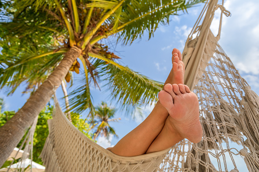 Relaxing beach vacation with woman's feet in hammock between coconut palm tree. Exotic tropical island hotel resort. Sunny warm day with blue sky.