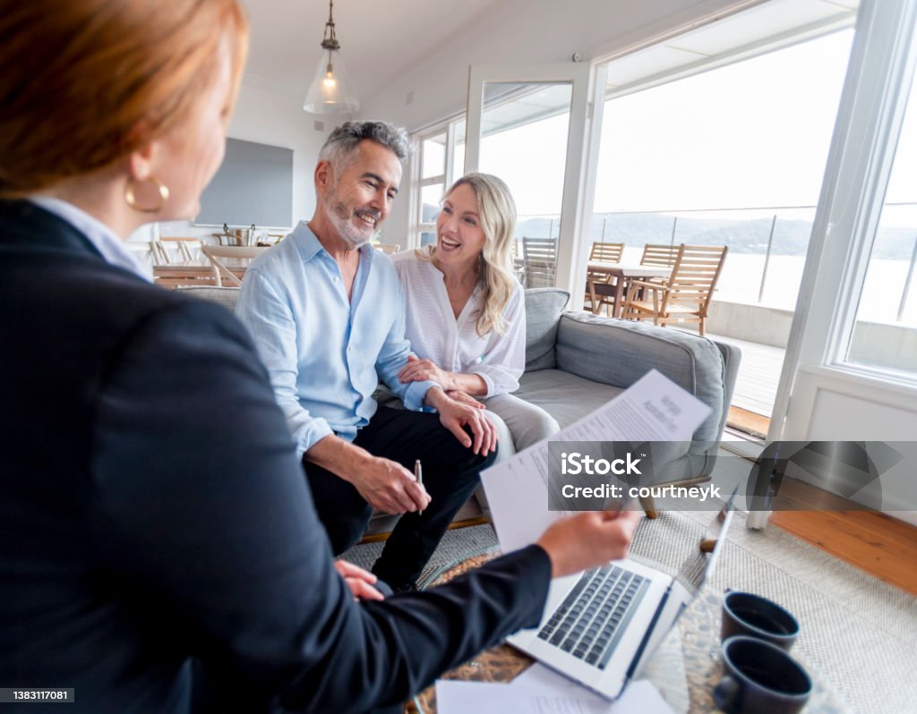 Happy mature couple meeting investments and financial advisor at home Happy mature couple meeting investments and financial advisor at home. They are happy and smiling sitting in the living room. The advisor is holding a document. There is a laptop on the table Real Estate Agent Stock Photo