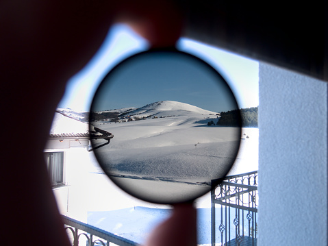 View of the snow-covered mountain peak through round tinted glass. The person holding camera ND filter and checking its opacity