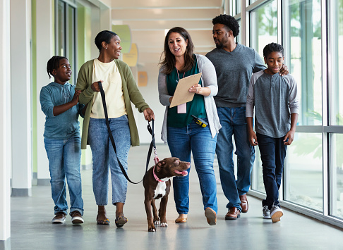 A volunteer at an animal shelter helping an African-American family with two boys adopt a rescue dog. They are walking down the hallway with the dog on a leash. The worker is holding a clipboard up so the mother can read the paperwork needed to finalize the pet adoption.