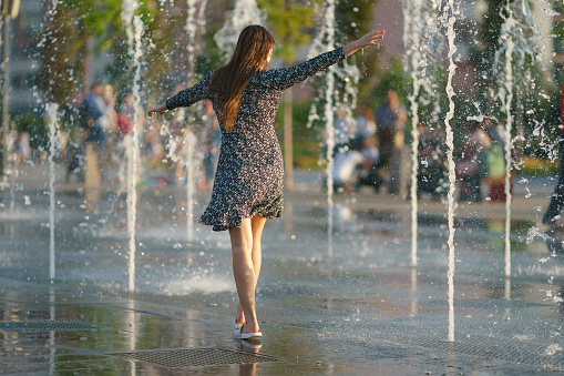 Moscow, Russia - June 19, 2021: Young woman playing with water of fountain in the Moscow public park. She is having fun and dancing. Hot summer amusement. Leisure concept. Back, rear view