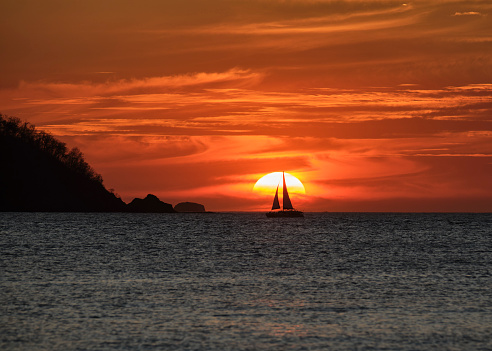 A spectacular orange sunset with swirling clouds and the silhouette of a sailboat in front of the sun in Potrero Costa Rica.