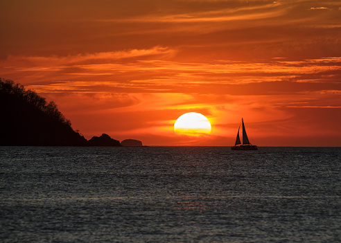 A spectacular orange sunset with swirling clouds and the silhouette of a sailboat sailing toward the sun in Potrero Costa Rica.