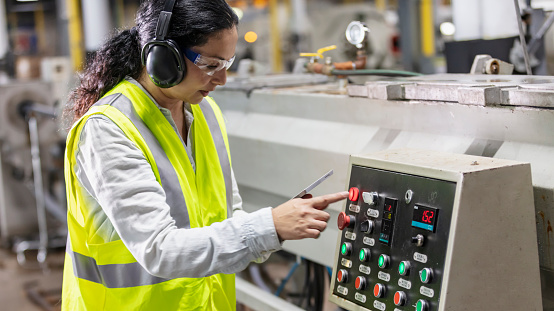 A mature Hispanic woman in her 40s working in a plastics factory, standing next to manufacturing equipment, at a control panel, finger about to push a button. She is wearing protective eyewear, ear protectors and a reflective vest, holding a walkie-talkie.