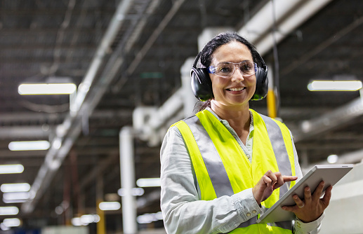 A mature Hispanic woman in her 40s working in a plastics factory, standing on the factory floor next to manufacturing equipment, using a digital tablet. She is wearing protective eyewear, ear protectors and a reflective vest, smiling and looking toward the camera.