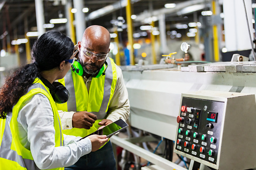 Two multi-ethnic workers working in a plastics factory, standing on the factory floor, at the control panel of one of the machines. The Hispanic woman is holding a digital tablet. She and her coworker, an African-American man, are conversing face to face.