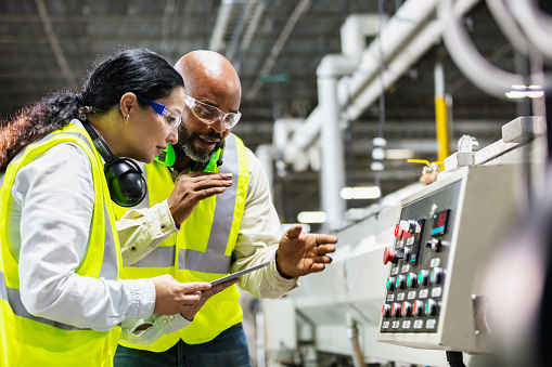 Two multi-ethnic workers working in a plastics factory, standing on the factory floor, looking at the control panel of one of the machines. The African-American man is pointing to the panel. His coworker, an Hispanic woman, is holding a digital tablet.