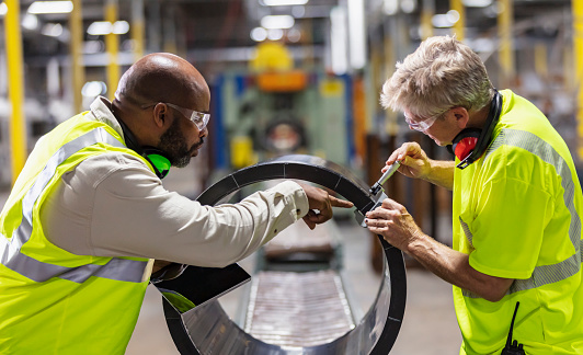 Two multiracial men working in a plastics factory. The African-American man, in his 50s, is the manager, holding a digital tablet and talking to the other man, a senior in his 60s, who is measuring a large plastic cylinder on a conveyor belt.