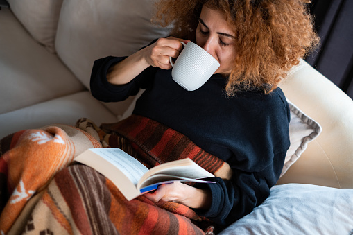 Young woman sitting on sofa with mug of coffee in reading a book.