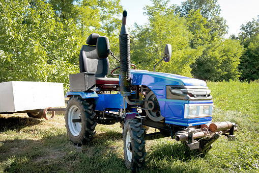 Small mini blue modern new tractor with trailer standing at farm, field, nature countryside during sunny summer day. Small agricultural machinery. Rural country farmland background.