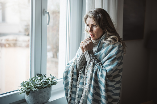Unwell mature woman in blanket sit in cold living room suffer from air conditioner lack. She is sitting next to radiator.No heating concept.