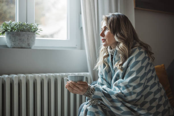 Unwell woman feel cold in home with no heating Unwell mature woman in blanket sit in cold living room suffer from air conditioner lack. She is sitting next to radiator.No heating concept. cold temperature stock pictures, royalty-free photos & images