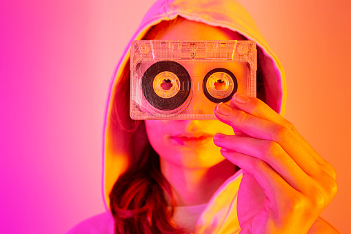 Teenager girl holding plastic transparent cassette tape. Old media - audio format technology. Bright pink and orange background, pop art style poster. Layout with free copy (text) space.