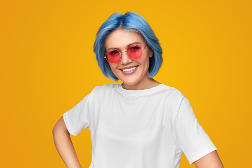Happy female hipster in white t shirt and trendy sunglasses holding hand on waist and smiling for camera against yellow background