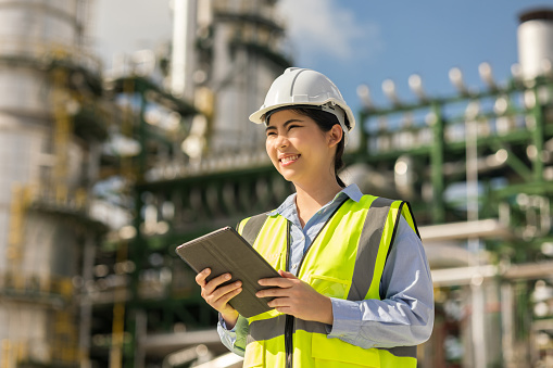 Asian engineer manager woman with white safety helmet standing front of oil refinery. Industry zone gas petrochemical. Factory oil storage tank and pipeline. Workers in the refinery construction.