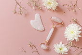 Rose quartz facial massage roller over pink background with gypsophila flowers. Massage tools with jade stone, anti-aging, anti-wrinkle beauty skincare tool. Top view flat lay. Copy space.