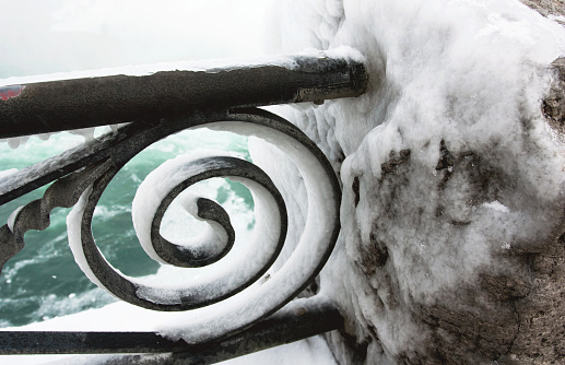 A snow and ice covered railing during winter at Niagara Falls. The surrounding mist causes water to freeze on any object nearby. Niagara’s waters can be seen in the background.