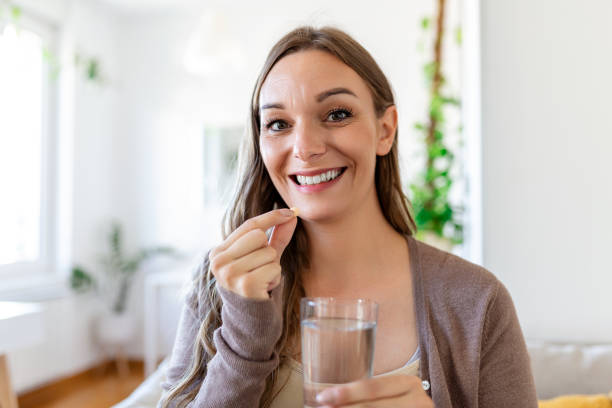 glückliche frau zu hause sitzt am tisch zu hause hält tasse wasser und pille coronavirus-behandlung nahaufnahme verschwommener hintergrund - vitamin enriched stock-fotos und bilder