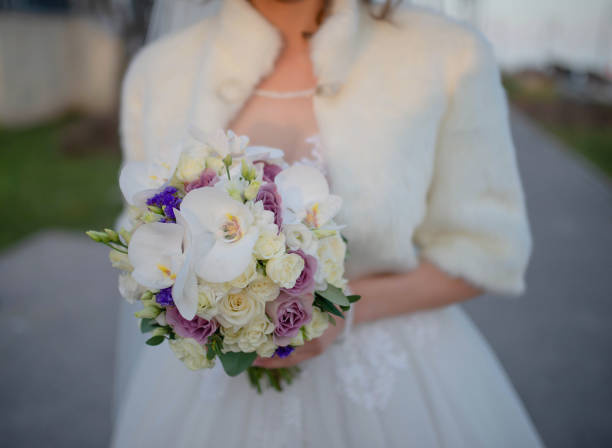 Bride wearing an exquisite white dress while holding an elegant flower bouquet featuring pastel colored roses and white orchids, concepts for romantic bridal vibes and happiness of getting married.