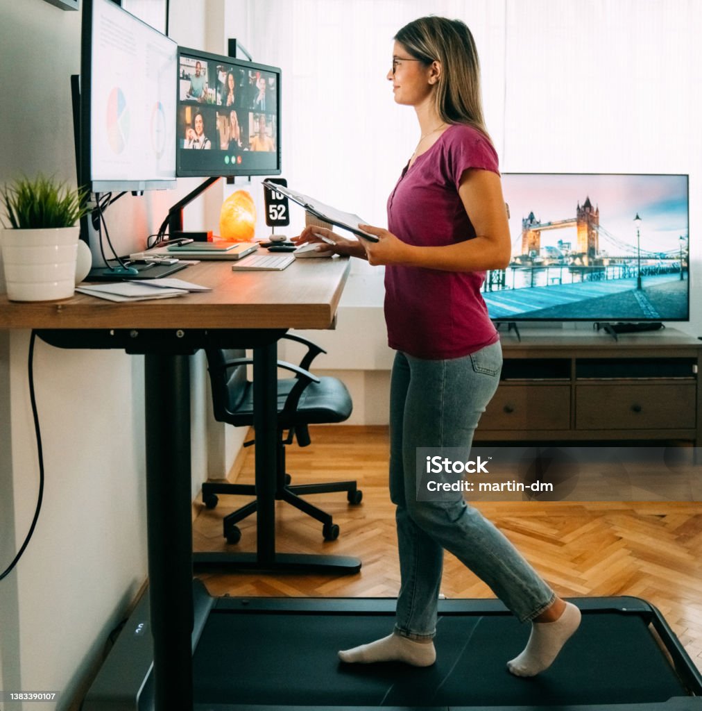 Woman at standing desk home office talking on business video call Woman working from home at standing desk is walking on under desk treadmill Ergonomics Stock Photo