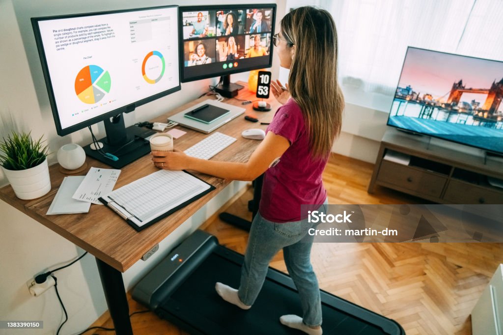 Woman at standing desk home office talking on business video call Woman working from home at standing desk is walking on under desk treadmill Working At Home Stock Photo