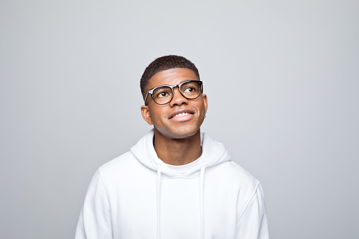 Pensive african young man wearing white hoodie and eyeglasses looking up and smiling. Studio portrait on grey background.