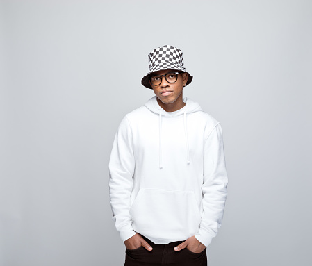 African young man wearing white hoodie, bucket hat and eyeglasses, standing with hands in pockets and looking at camera. Studio portrait on grey background.