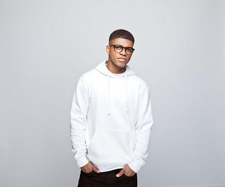 African young man wearing white hoodie and eyeglasses, standing with hands in pockets and looking at camera. Studio portrait on grey background.