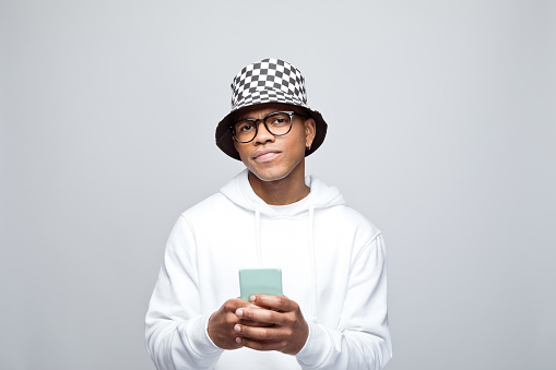 Handsome african young man wearing white hoodie, bucket hat and eyeglasses, holding smart phone in hands and looking at camera. Studio portrait on grey background.