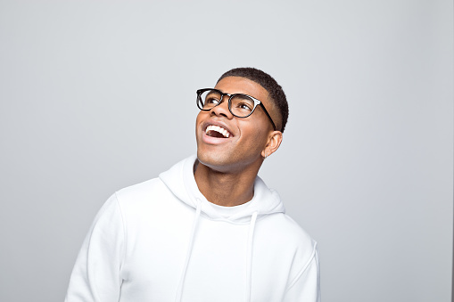 Surprised african young man wearing white hoodie and eyeglasses, looking away with mouth open. Studio portrait on grey background.