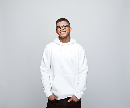 Happy african young man wearing white hoodie and eyeglasses, standing with hands in pockets and smiling at camera. Studio portrait on grey background.