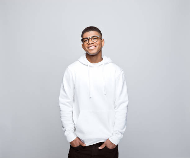 Portrait of friendly young man Happy african young man wearing white hoodie and eyeglasses, standing with hands in pockets and smiling at camera. Studio portrait on grey background. hooded-shirt stock pictures, royalty-free photos & images