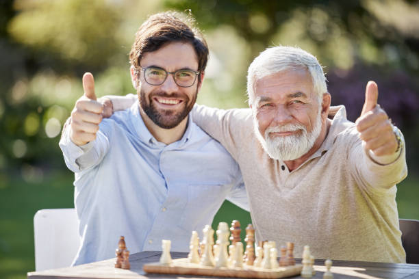 tiro de um jovem e idoso jogando xadrez juntos e mostrando polegares para fora - board game success victory winning - fotografias e filmes do acervo