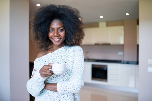 istock Smiling african woman drinking coffee at home, looking at camera. 1383426110