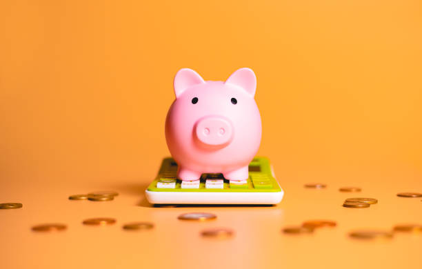 A piggy bank over a green calculator with scattered coins isolated on orange background in studio photography. Concepts of saving money, finance and investments. A piggy bank over a green calculator with scattered coins isolated on orange background in studio photography. Concepts of saving money, finance and investments. ira gold companies stock pictures, royalty-free photos & images