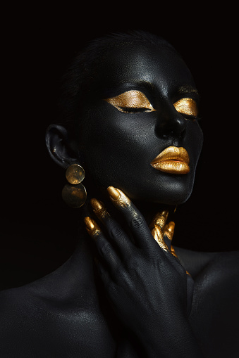 Beauty woman painted in black skin color body art, gold makeup lips eyelids, fingertips nails in gold color paint. Professional gold makeup