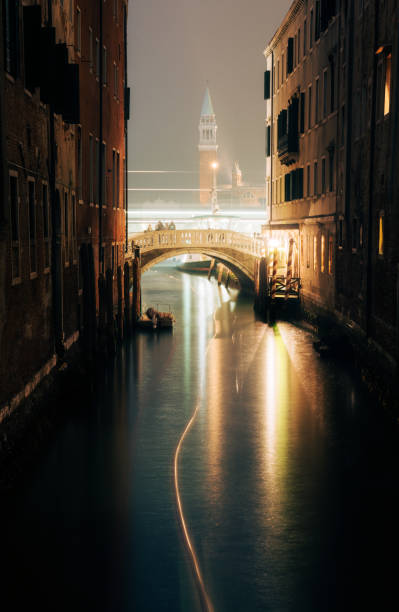 Night time in Venice and light trails from a small boat manoeuvre up a side canal towards the city's Grand Canal. In the distance we see the iconic shape of the Church of San Giorgio Maggiore.