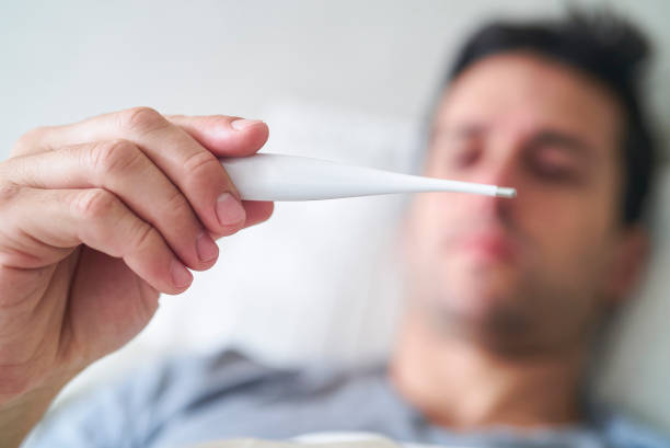 Close-up shot with selective focus of hispanic adult man taking temperature with digital thermometer while lying in bed during daytime
