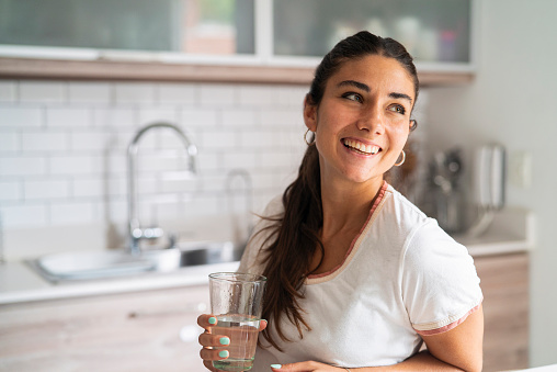 Front shot of beautiful smiling young adult hispanic woman holding glass of water while looking up standing in kitchen during daytime