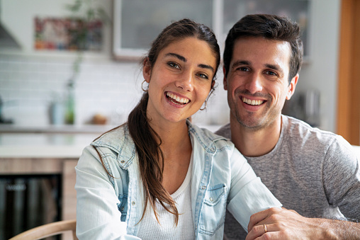 Front shot of hispanic young adult couple sitting in kitchen while smiling and looking at the camera during daytime
