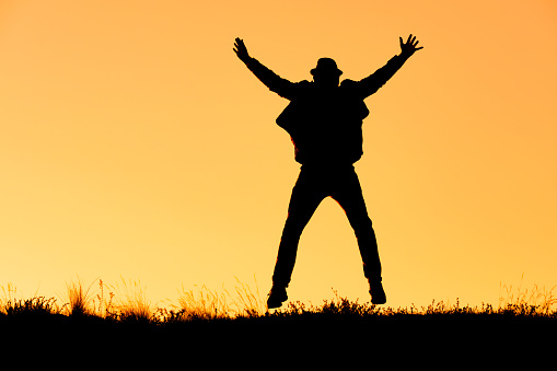 silhouette of a man jumping for joy in a hat, arms and legs in different directions. the background is orange, joy and fun in nature, a splash of emotions