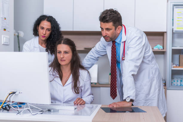 istock Two nurses ( female physicians ) and a male doctor standing next to a pc, mac ( desktop computer ) in the doctor's office while discussing a medical report. 1383478507