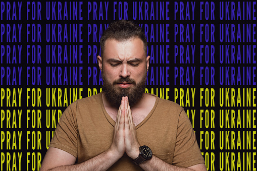 Portrait of Ukrainian man praying for Ukrainian safety and peace isolated over blue and yellow flag with lettering Pray for Ukraine.