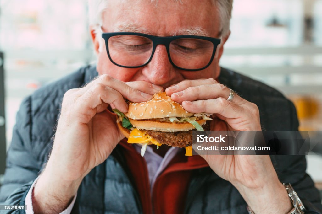 Senior man eating burger in fast food restaurant Close up color image depicting a senior man wearing spectacles eating a freshly cooked burger in a fast food restaurant. The burger is messy, with lettuce and cheese hanging out of the bun. Room for copy space. Eating Stock Photo