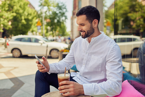 Young man using smartphone and drinking coffee outside a coffee shop in the city