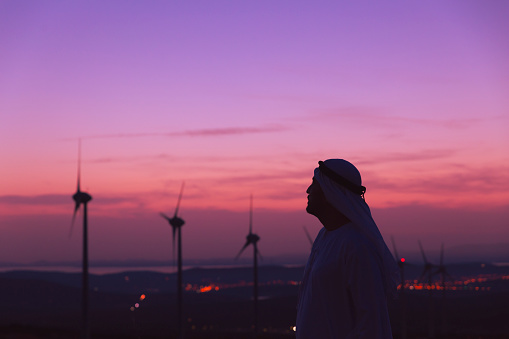 Arab Sheikh with traditional Emirati clothes looking for the energy industry and standing beside a wind turbines farm power station at sunset time.