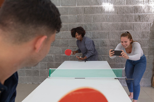 A male and a black female business executives  are playing ping-pong at a  leisure room while a female executive standing on one side of the table happily takes photos with her smartphone
