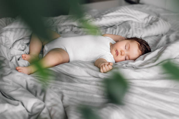 ベッド毛布の上に寝るの赤ちゃんの肖像画、前景にぼやけた緑の植物。寝室で幼児の子供を眠って、甘い夢を見てください。ソフトフォーカス、フリーコピースペース。育児と健康的な睡眠� ストックフォト