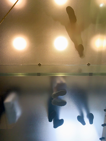 Directly below view of a group of people standing on a glass floor with spotlights shining to show only silhouettes of legs and feet.