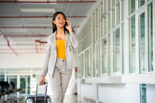Businesswoman Dragging suitcase luggage bag, walking to passenger boarding in Airport. Woman travel to work. Asian tourist female wearing suits and using smartphone. Businesswoman Travel concept